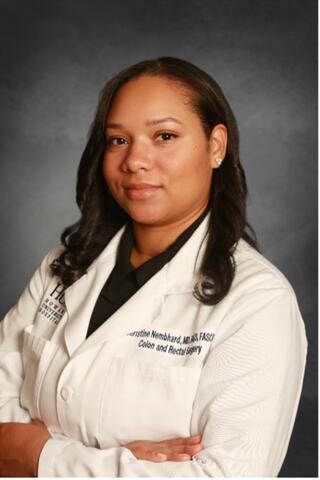 Photo of Dr. Christine Nembhard wearing a white lab Howard University coat that also says 'Christine Nembhard, MBBS, FACS, FASCRS Colon and Rectal Surgery'. She smiles with her arms crossed against a dark gray background.